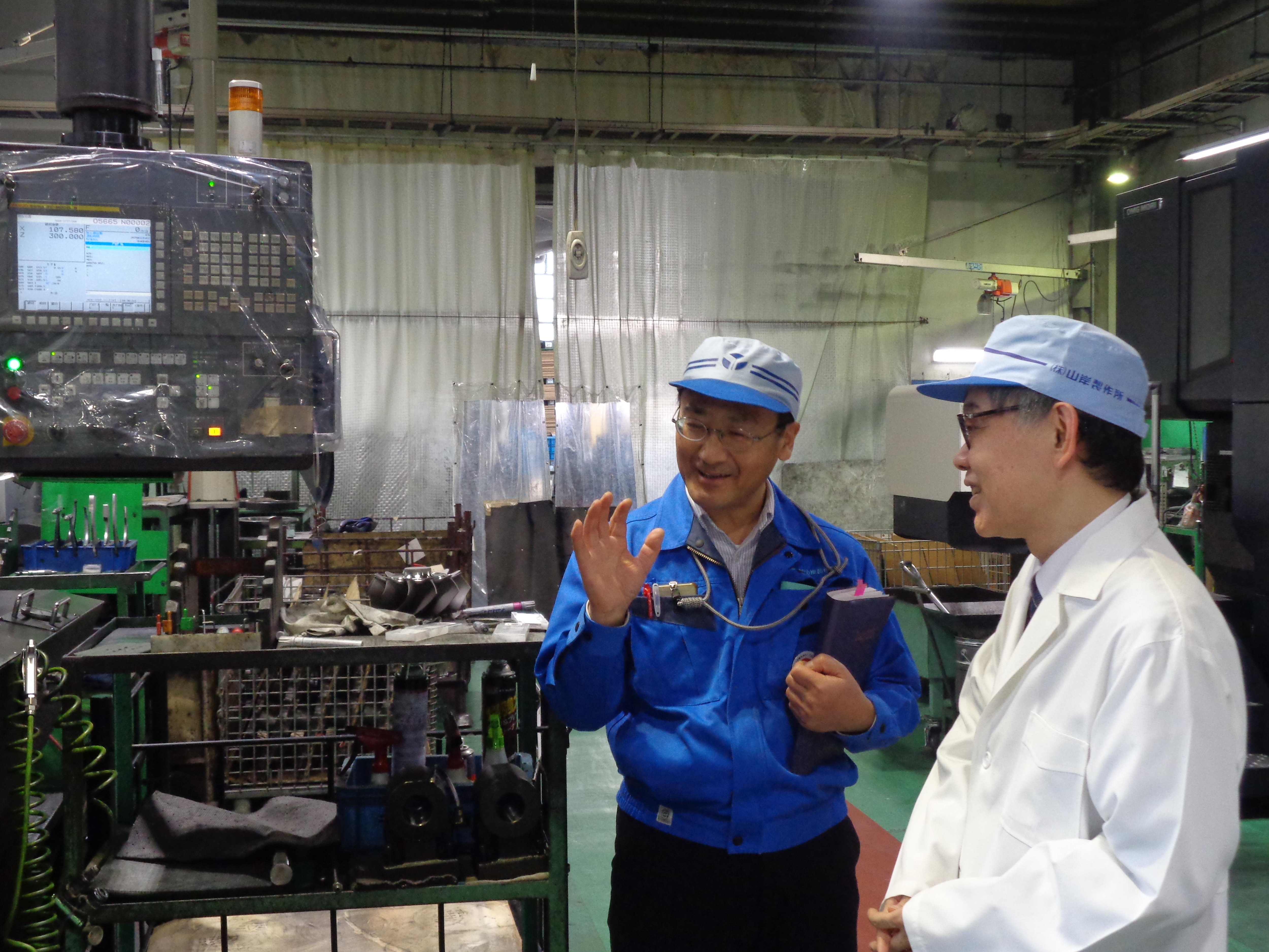 株式会社山岸製作所の機械の前で山岸代表取締役社長から説明を受ける岩成事務総長の様子